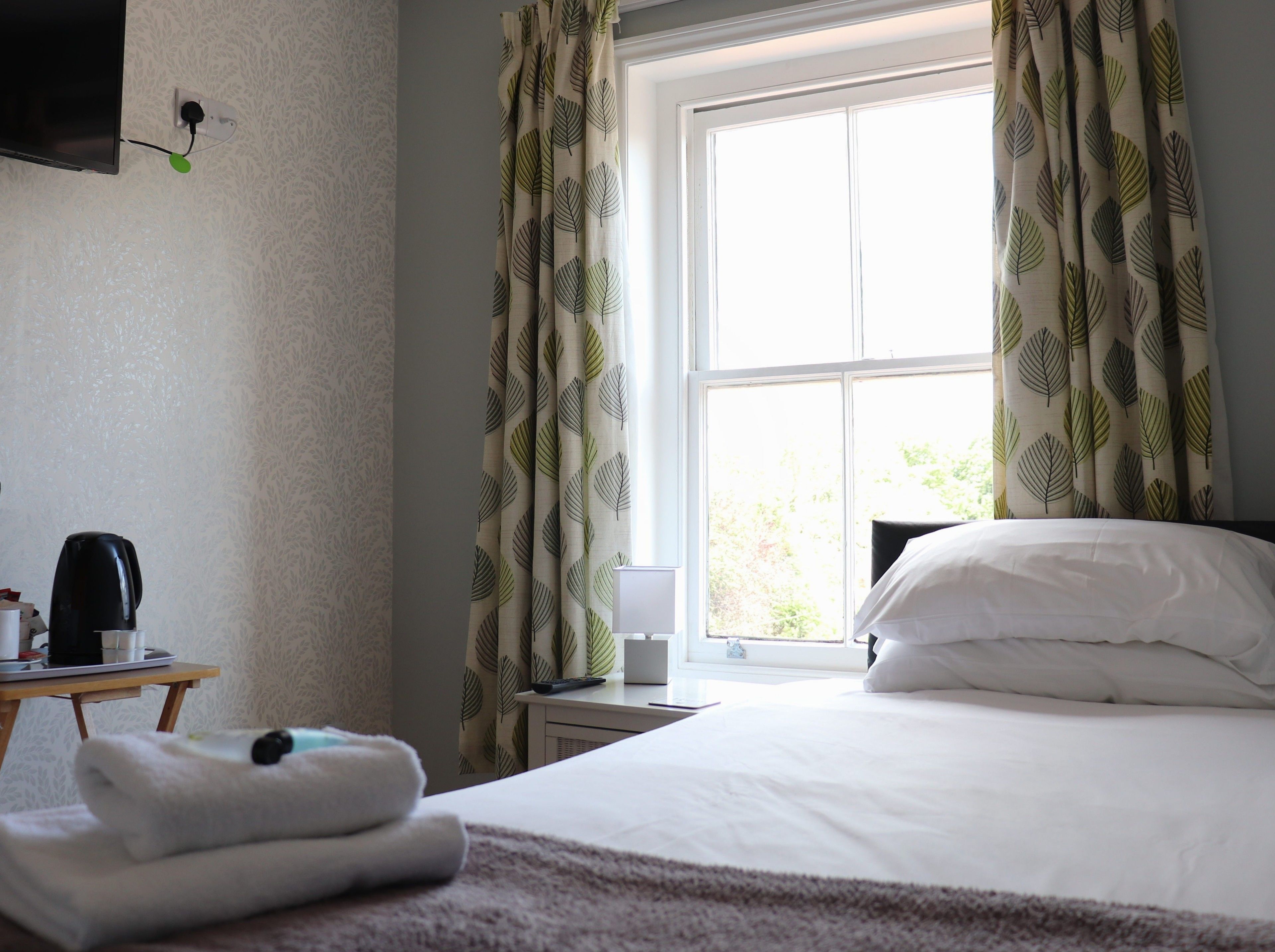 Habitación de hotel con cama, mesita de noche con hervidor, lámpara y ventana con cortinas de diseño floral.