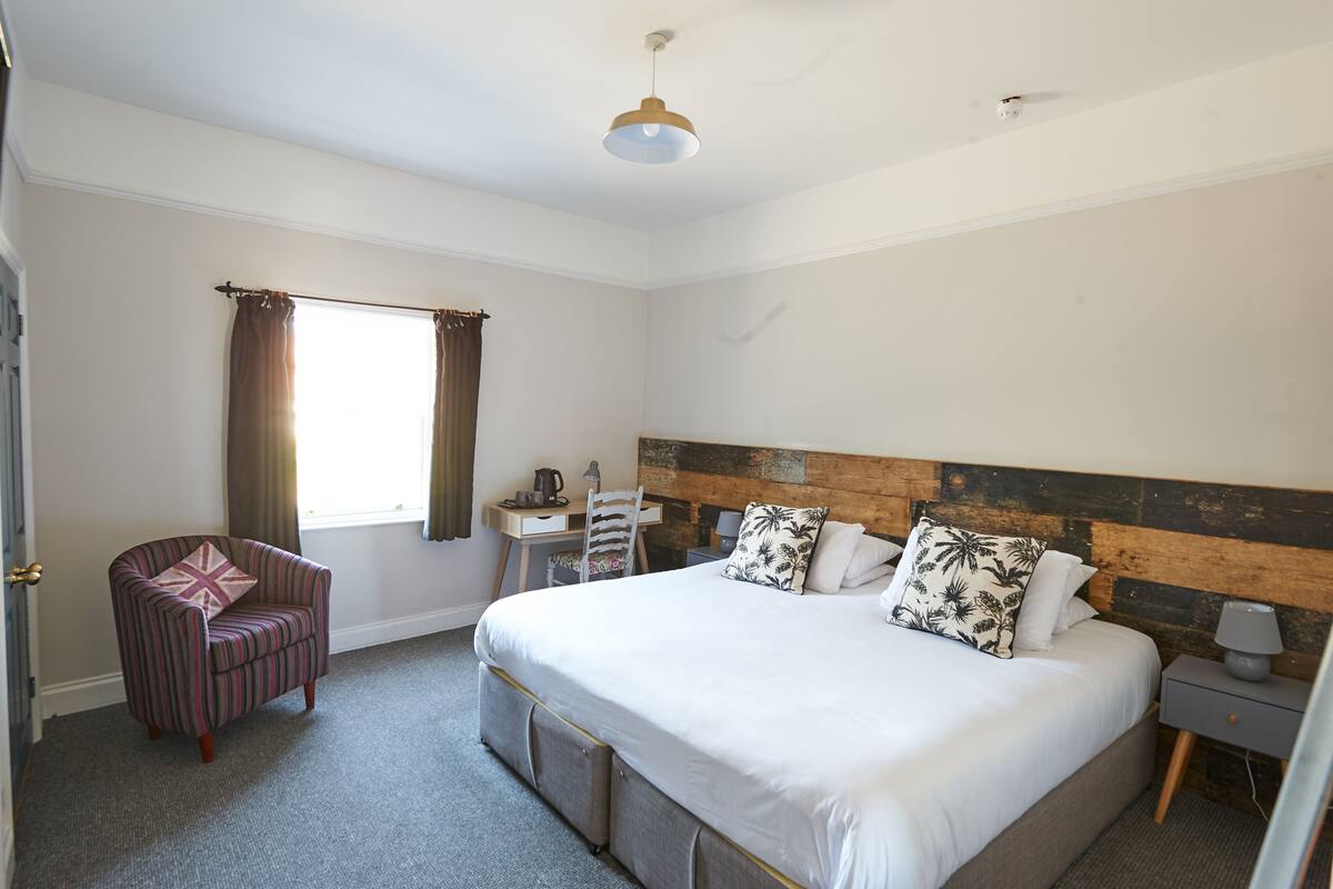 A bedroom with a large bed, a chair, a desk, and a window with curtains.