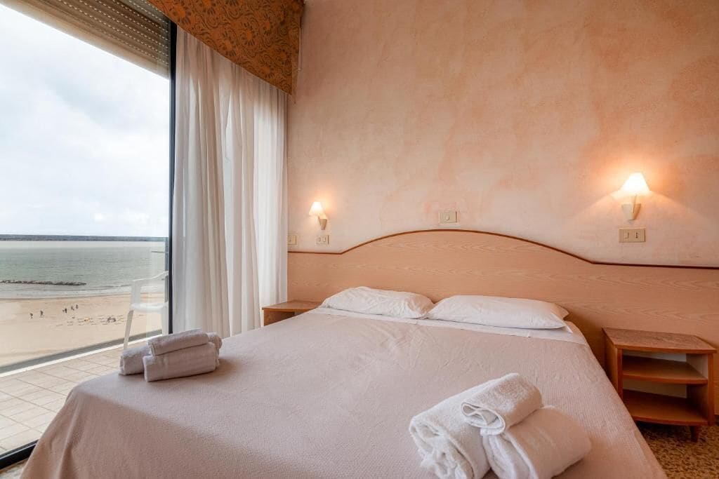 Une chambre d’hôtel comprenant un lit, deux tables de chevet, une fenêtre donnant sur la plage et des lampes fixées au mur.