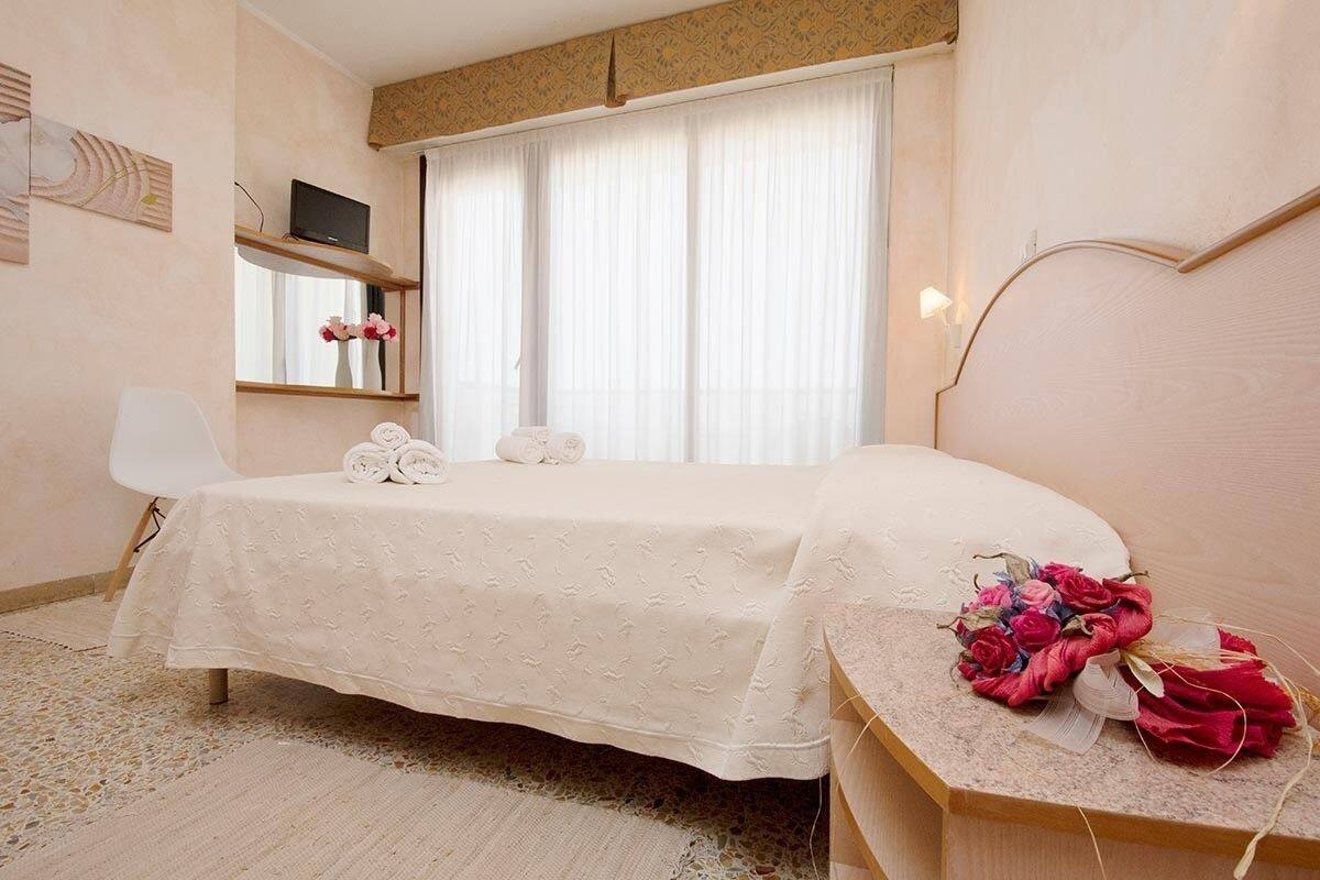 Un dormitorio con cama, una silla, una mesita de noche con un ramo de flores y un televisor en un estante.