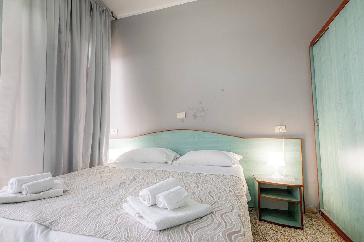 Habitación de hotel con una cama, dos almohadas, una mesita de noche con lámpara y una toalla doblada sobre la cama.