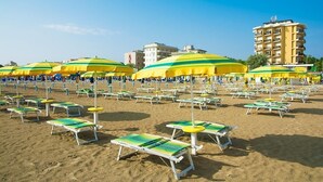 A beach with rows of sun loungers and umbrellas, a clear sky, and buildings in the background.