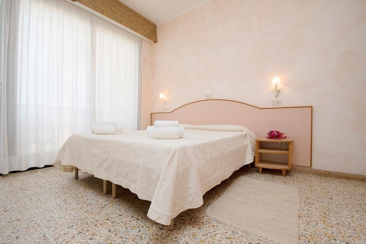Un dormitorio con cama, dos lámparas de noche, una mesita de noche de madera y una ventana con cortinas de gasa.