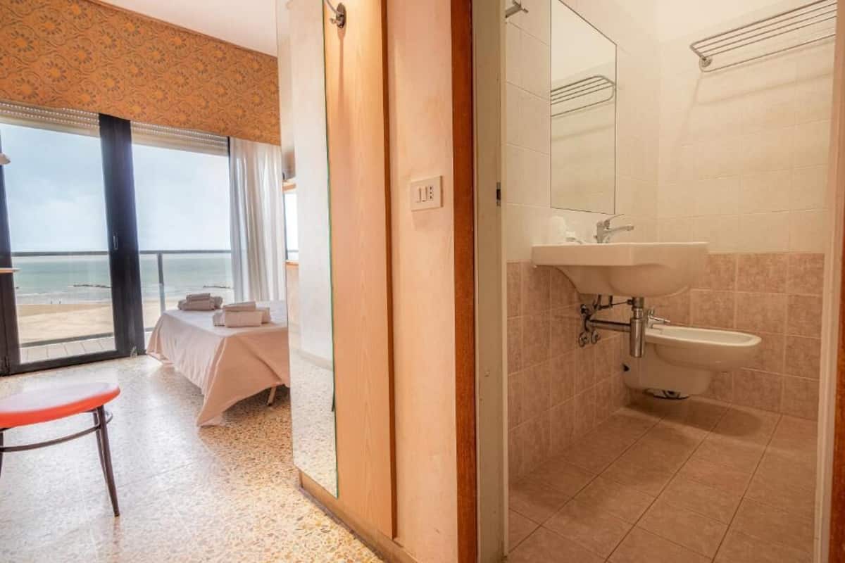 Habitación de hotel con cama, una silla, baño con lavamanos y espejo, y vista a la playa.