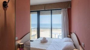 A bedroom with a bed, bedside tables, a nightstand, a window with a view of the beach, and a door.