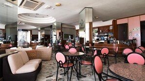 A restaurant with round tables and cushioned chairs, a stone floor, and a bar area with shelves of bottles.