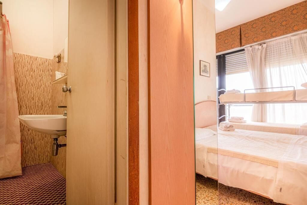 Una stanza con un letto, una finestra con tende, un lavandino e uno specchio.
