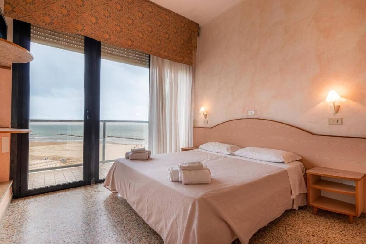 Habitación de hotel con cama, mesitas de noche, vista a la playa y un ventanal grande.