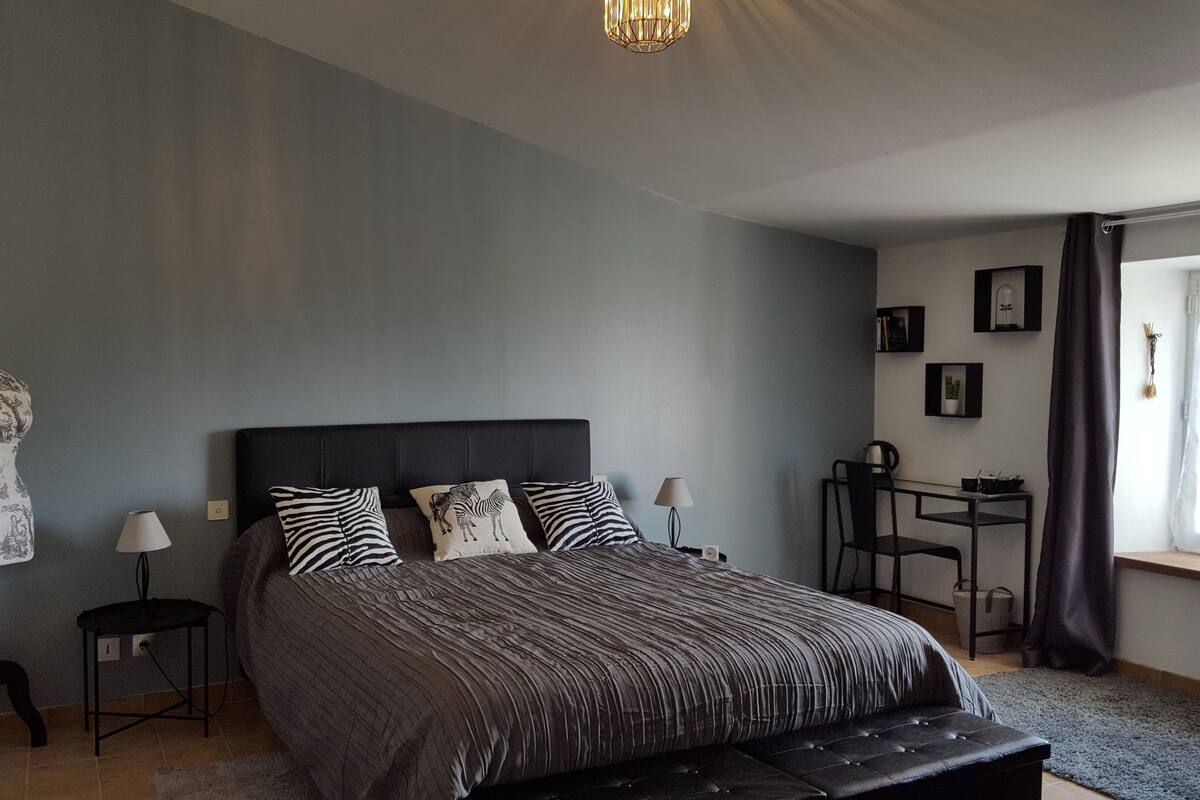 Ein Schlafzimmer mit einem großen Bett, zwei Nachttischen, einer Kommode, einem Fenster mit Vorhängen und einem Wandregal mit Büchern und einer Lampe.