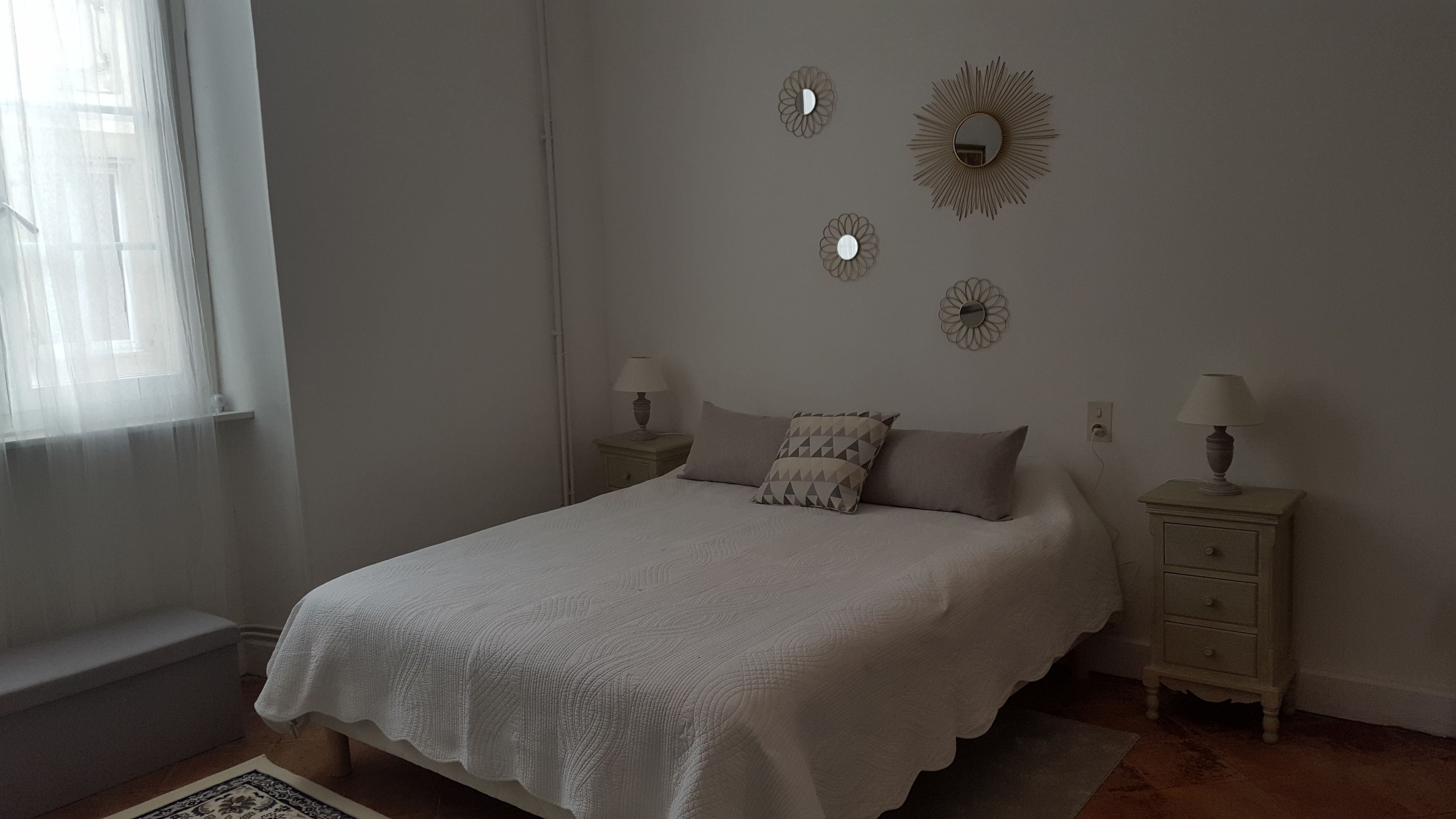 A bedroom with a bed, two bedside tables with lamps, a window with sheer curtains, and wall decor.