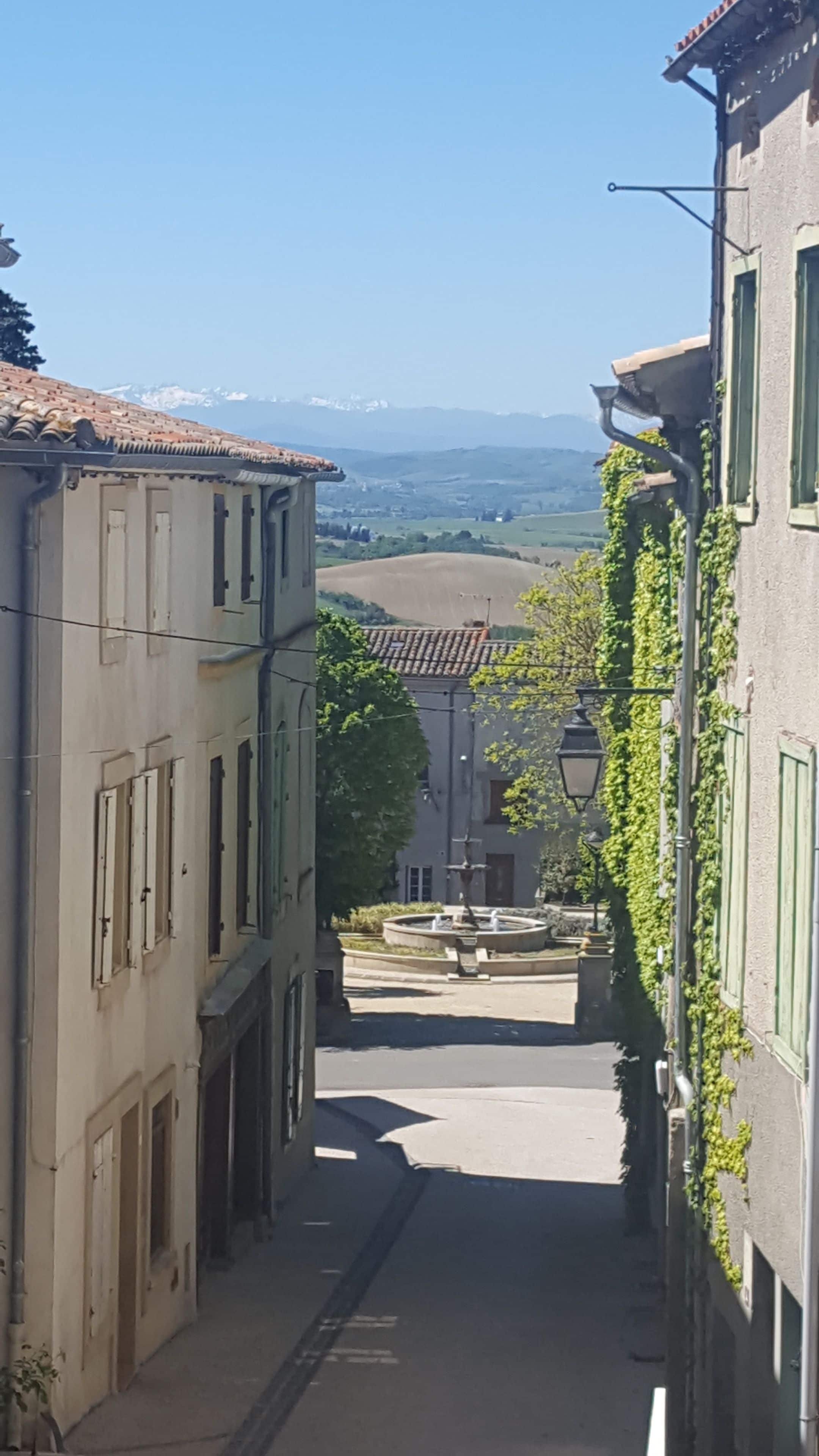 A narrow street flanked by buildings with a fountain in the distance.