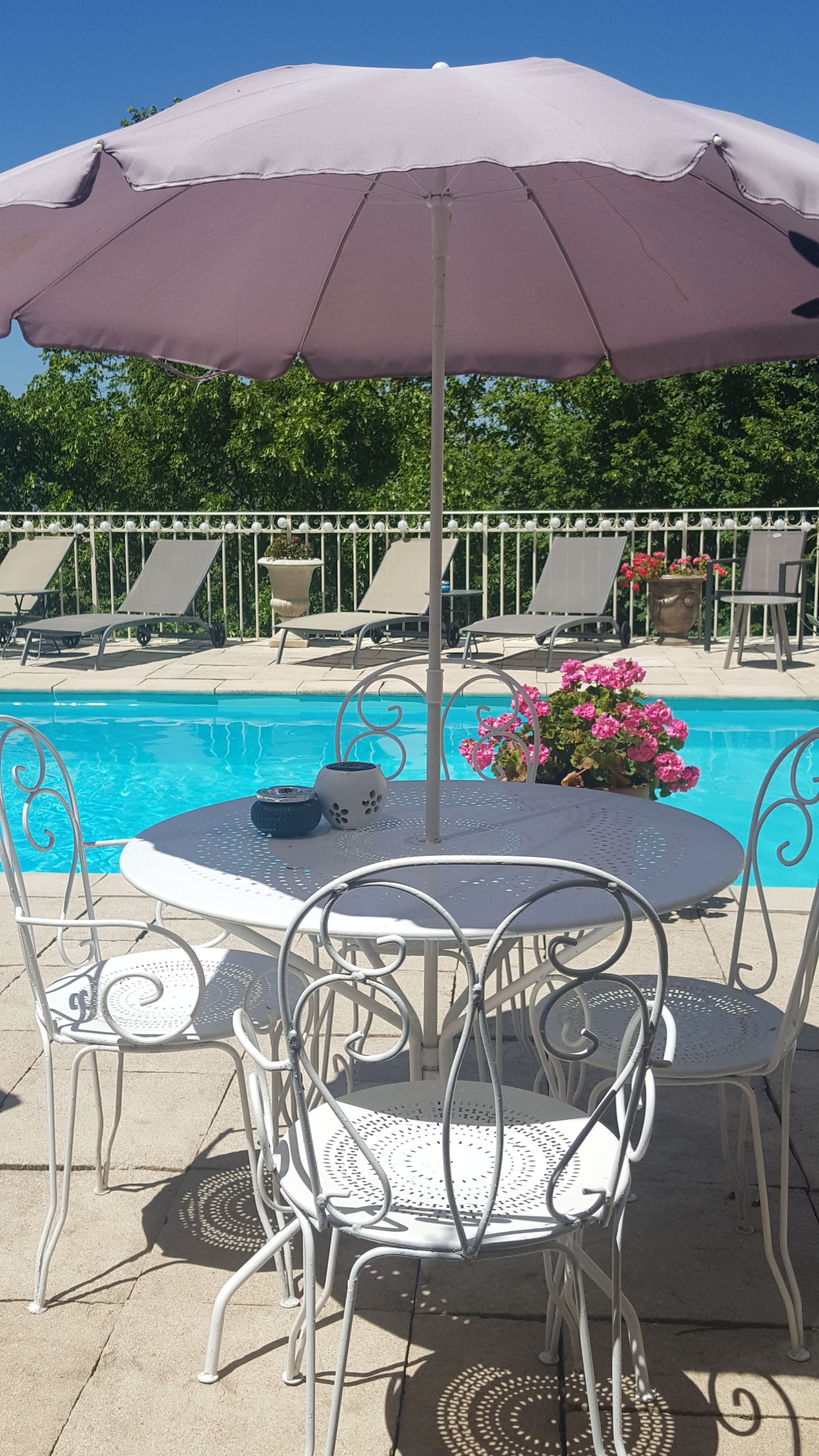 Área de piscina com mesa e cadeiras sob um grande guarda-sol, rodeada por espreguiçadeiras e piscina.