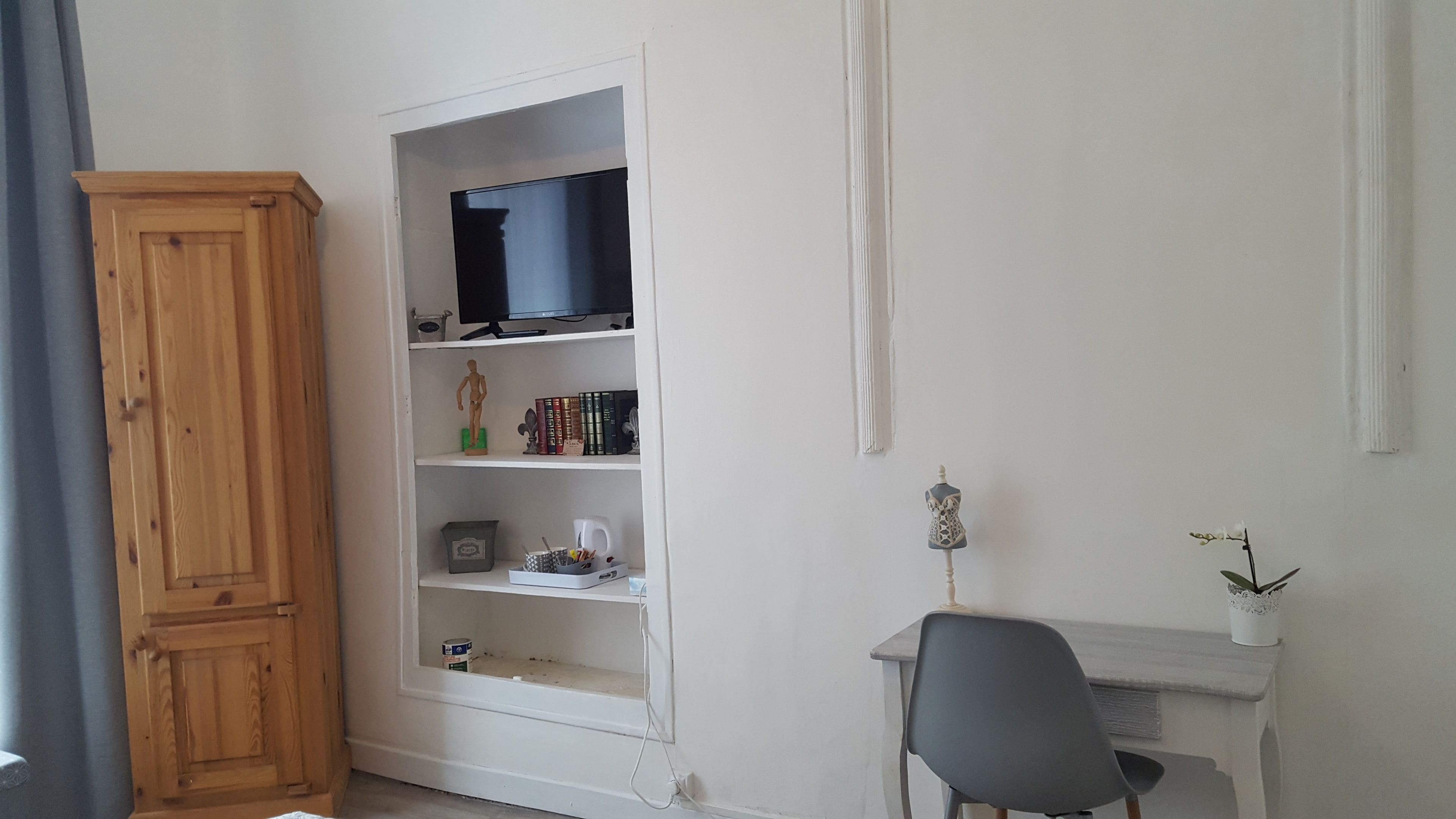 A room with a wooden wardrobe, a white desk with a chair, a television on a shelf, and a small plant on the desk.