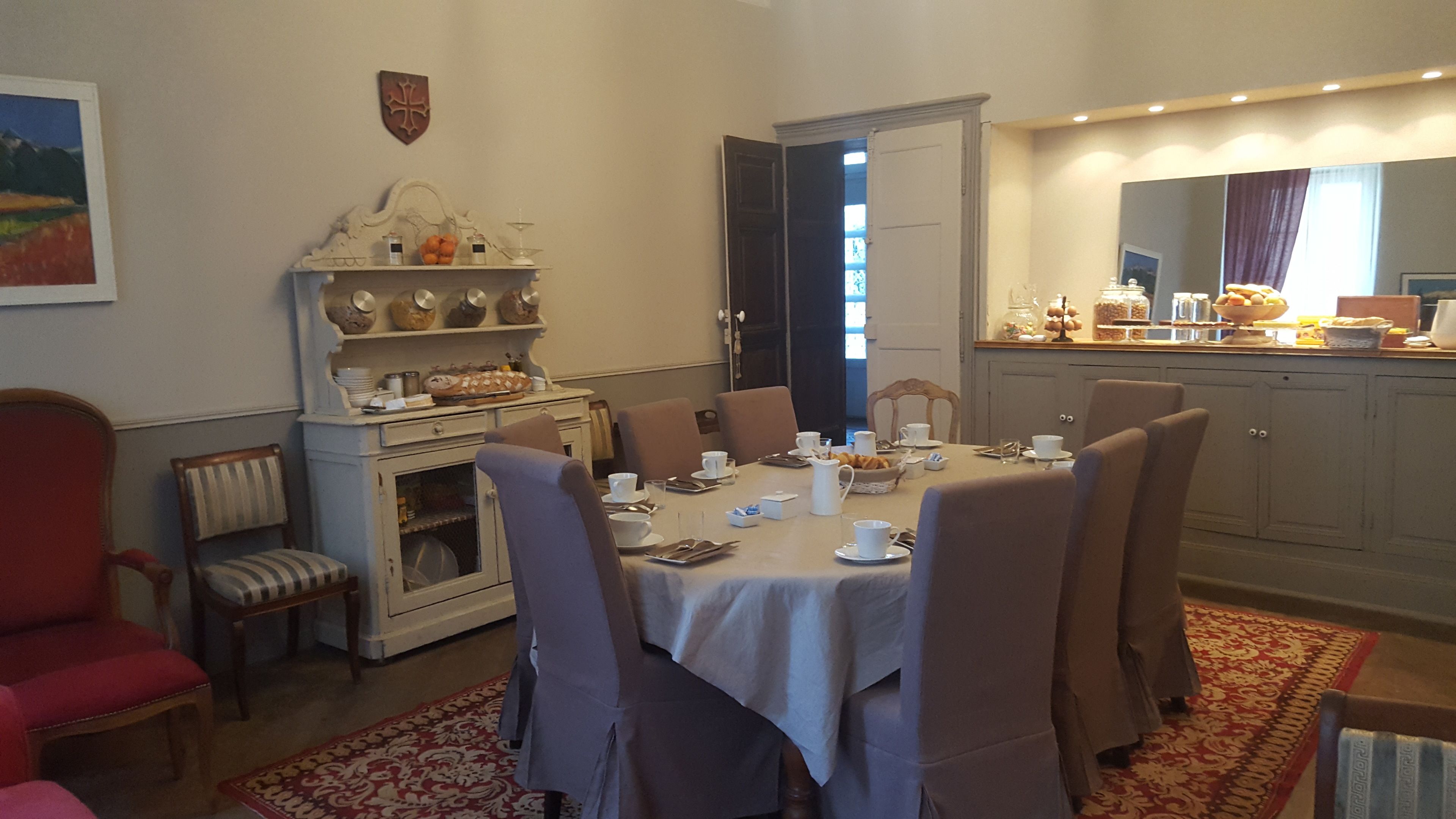 A dining area with a table set for four, surrounded by chairs. There is a buffet counter with various food items and a display of baked goods. The room has a traditional decor with a coat of arms on the wall and a red patterned rug on the floor.