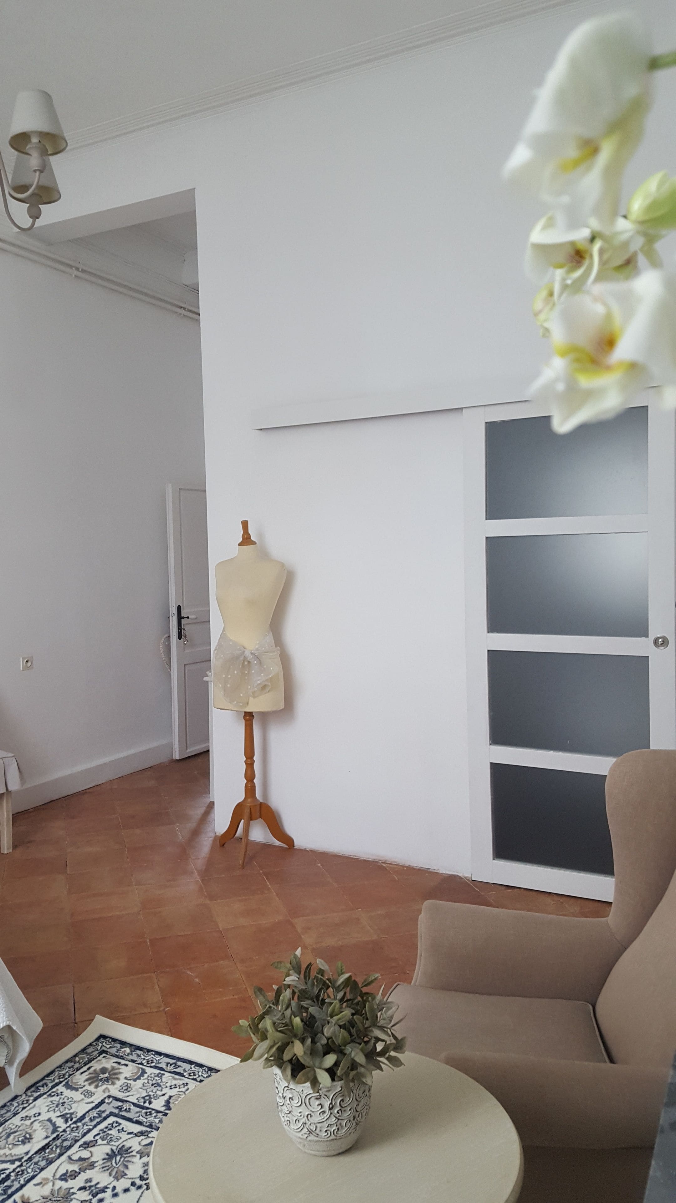 Un salon avec un mannequin sur un présentoir, un fauteuil beige, un tapis à motifs et une plante en pot posée sur une table ronde.