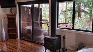 A room with a large window, a wood-burning stove, a bathtub, and a sliding glass door leading to an outdoor area. - Alaya Ascend  |  Legian Executive Pool-view Room |  Sunshine Coast Retreat (Eudlo)