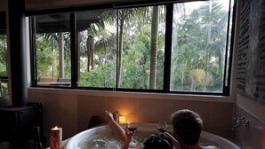 A person relaxing in a hot tub with a view of tropical foliage through large windows. - Alaya Ascend  |  Legian Executive Pool-view Room |  Sunshine Coast Retreat (Eudlo)