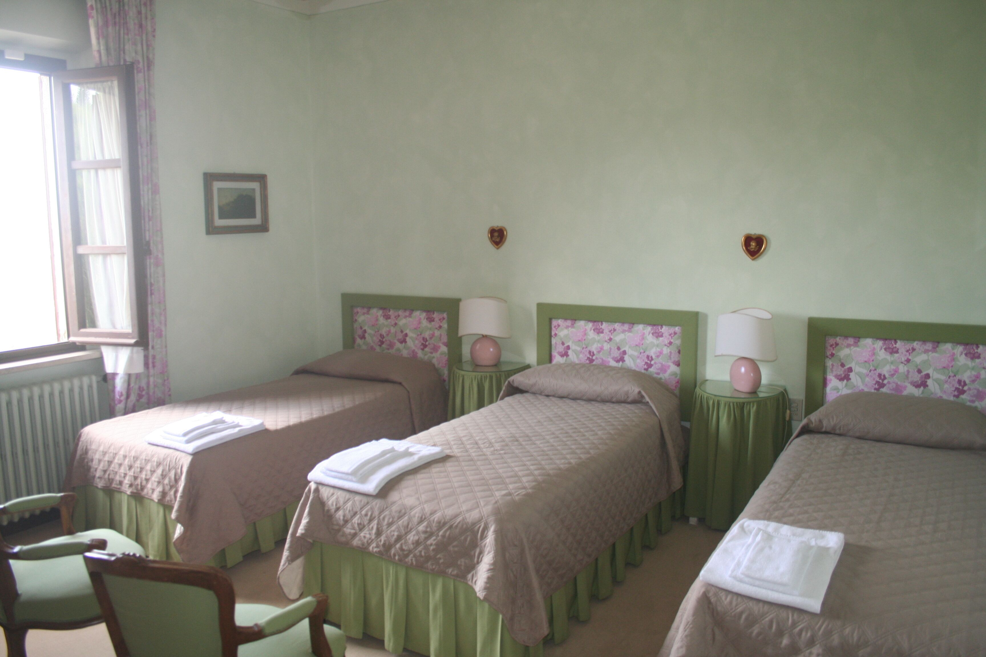A hotel room with two beds, each with a green headboard and floral wallpaper. There are two bedside tables with lamps, a window with curtains, and a chair.