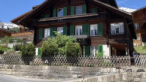 A traditional wooden chalet with multiple windows and balconies, surrounded by a stone wall and a wooden fence.