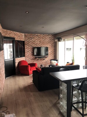 Ein modernes Wohnzimmer mit Backsteinwand, einem Flachbildfernseher, einem roten Sessel, einem schwarzen Sofa und einem Esstisch mit Stühlen.