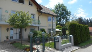 A two-story building with a balcony, a garden with a tree, and a metal fence.