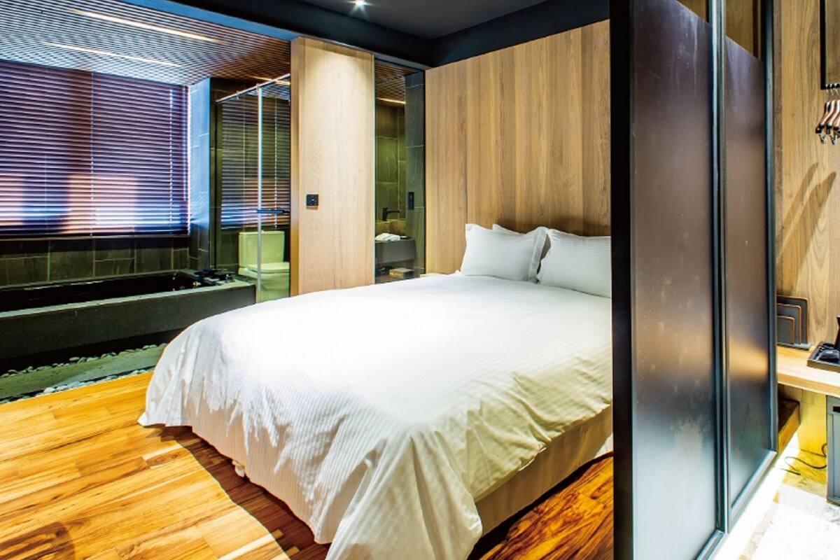 A modern hotel room with a large bed, wooden flooring, a glass shower enclosure, and a small kitchenette.