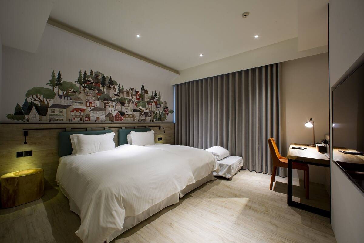 A hotel room with a large bed, a desk with a chair, a wall mural, and a curtain.