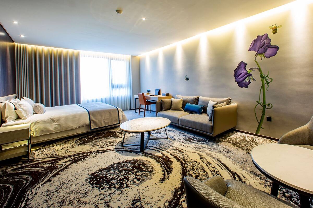 A modern hotel room with a bed, sofa, armchair, and a dining table.