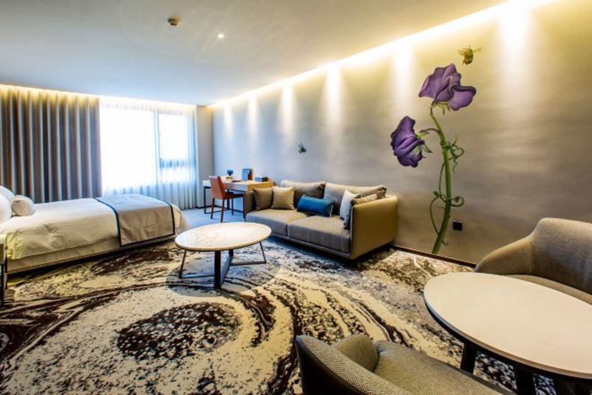 A modern hotel room with a sofa, two beds, a dining table, and a decorative wall with flowers.