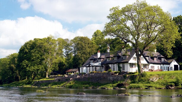 A row of white buildings with black roofs by a river, surrounded by trees. - Pwll-y-Faedda (Builth Wells)