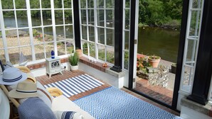 A sunroom with large windows overlooking a river, furnished with wicker chairs and a patterned rug. - Pwll-y-Faedda (Builth Wells)
