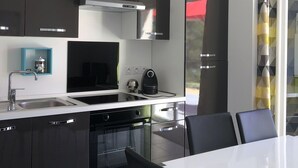 Une cuisine moderne avec des meubles noirs, un plan de travail blanc et un four encastré. On y trouve une table à manger avec deux chaises et une fenêtre agrémentée d’un rideau à motifs géométriques.