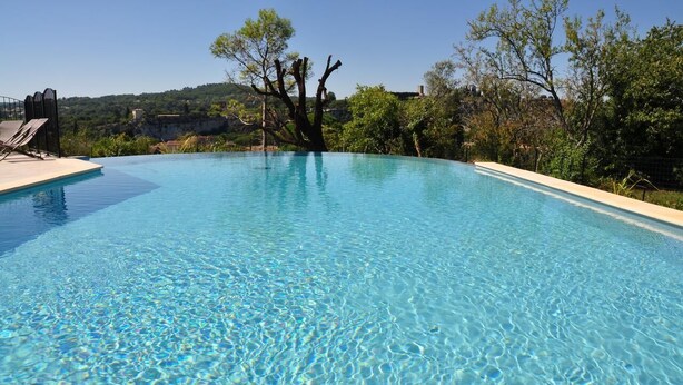 Une piscine aux eaux cristallines, dotée d’une terrasse en bois et de chaises longues, entourée d’arbres et bordée d’un paysage vallonné.