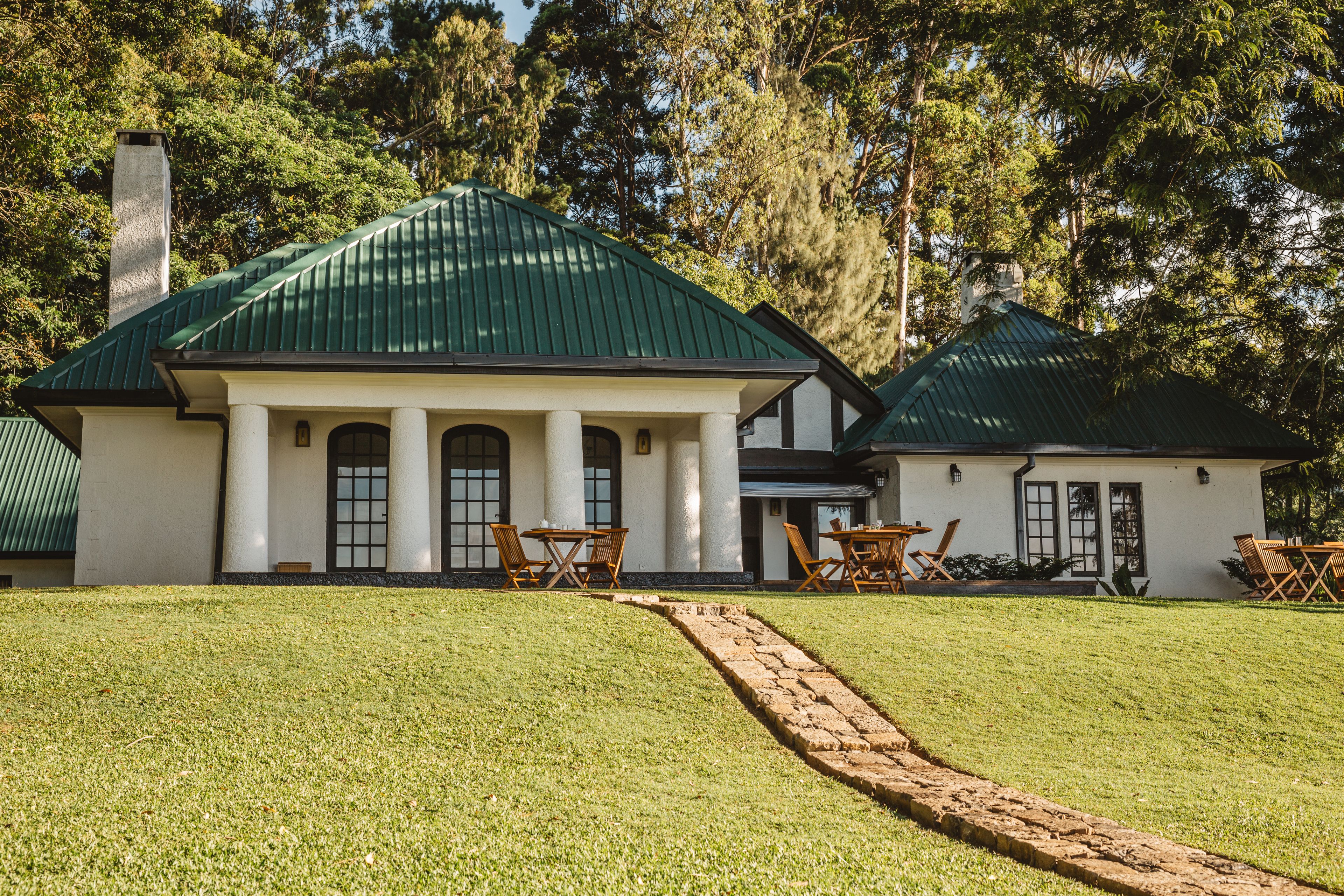 A large house with a green roof, white columns, and a paved driveway leading to a patio area with outdoor furniture.