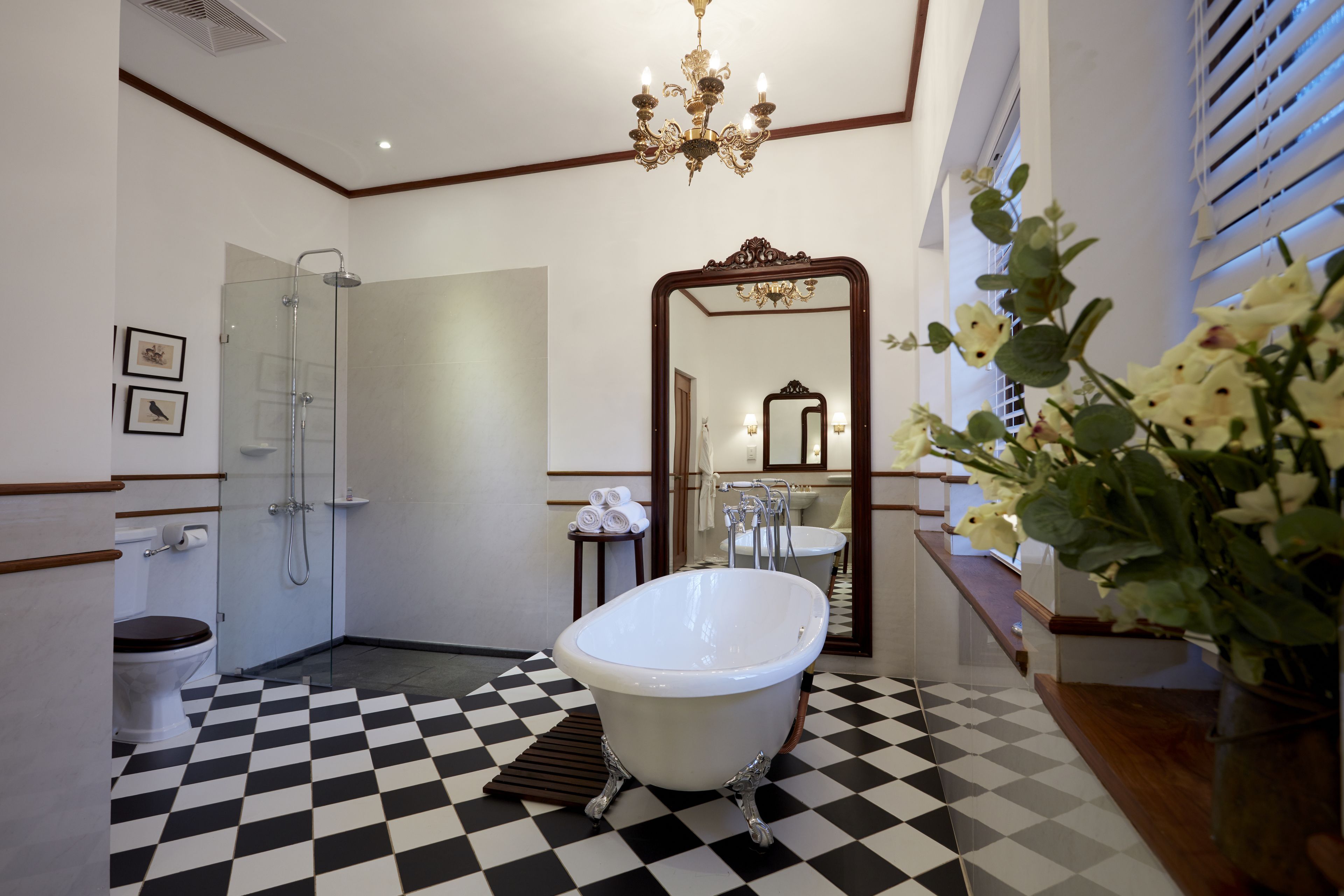 Une salle de bain avec un carrelage noir et blanc à motifs de damier, une baignoire sur pieds, une douche à parois de verre, un grand miroir et un lustre.