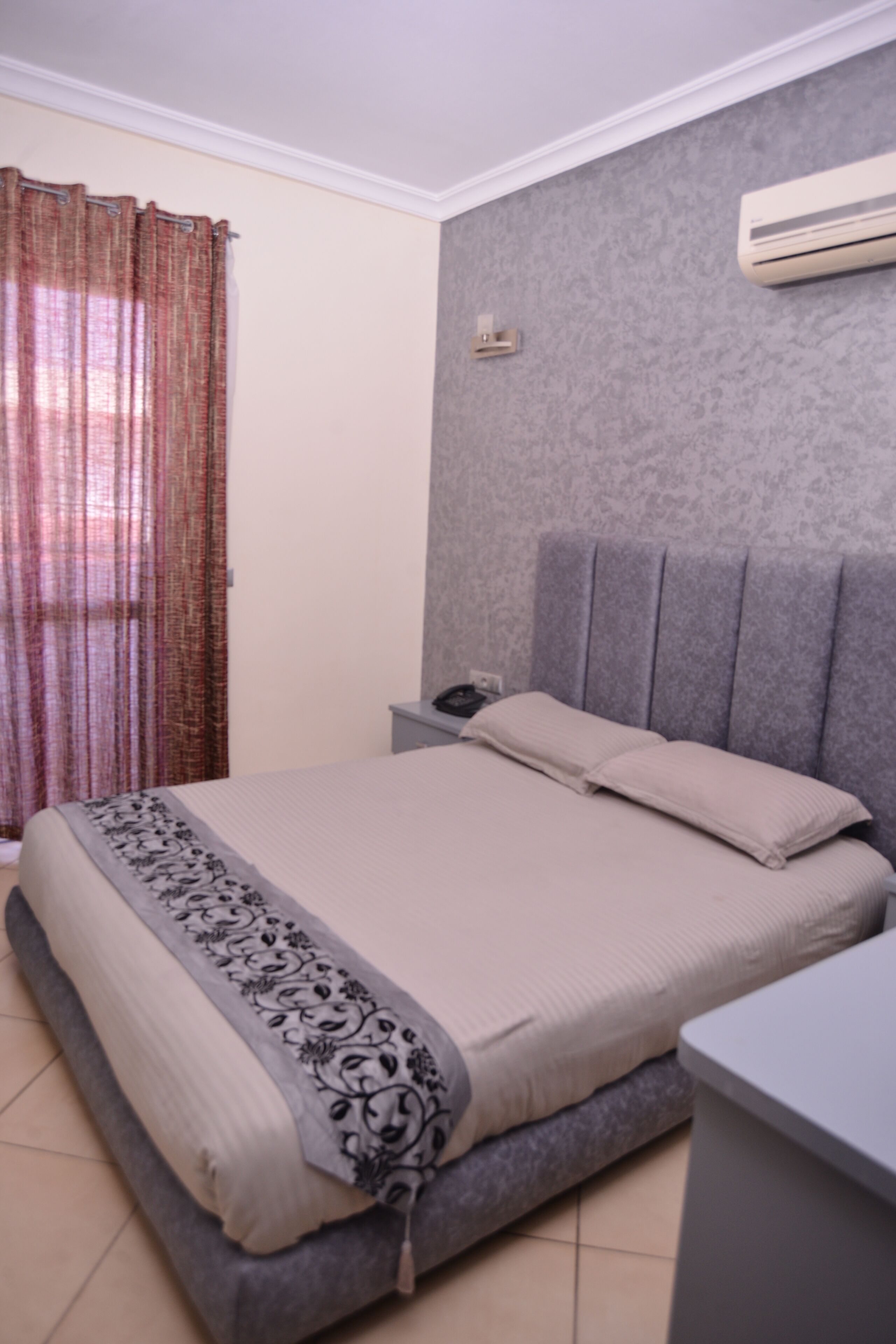 A bedroom with a bed, a bedside table, an air conditioning unit, and a curtain.