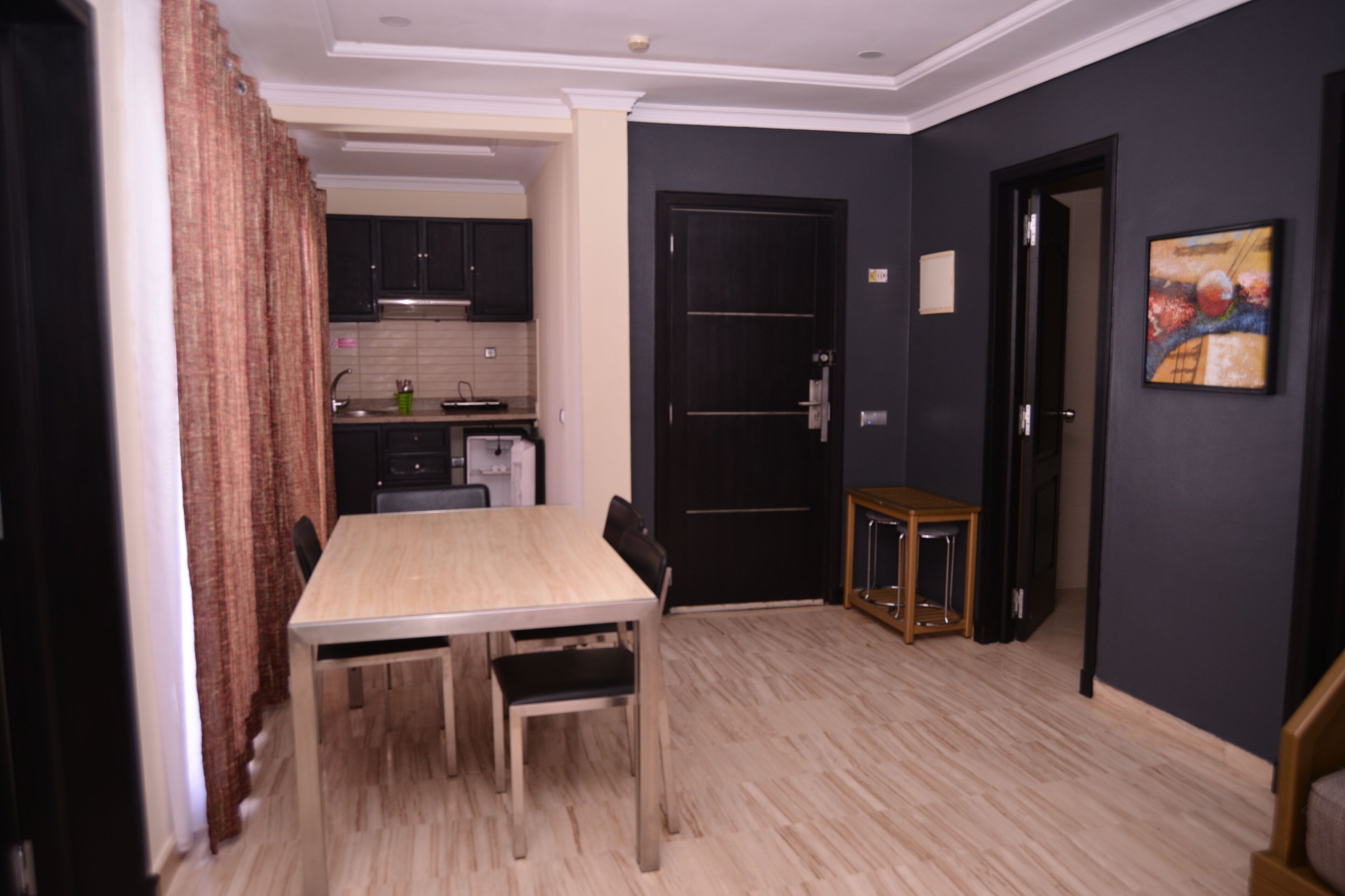 A dining area with a table and chairs, a kitchen in the background, and a dark-colored wall with a painting.