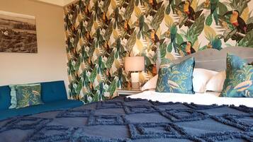 A bedroom with a bed, blue and white pillows, a nightstand with a lamp, and a wall with a tropical leaf and bird pattern.
