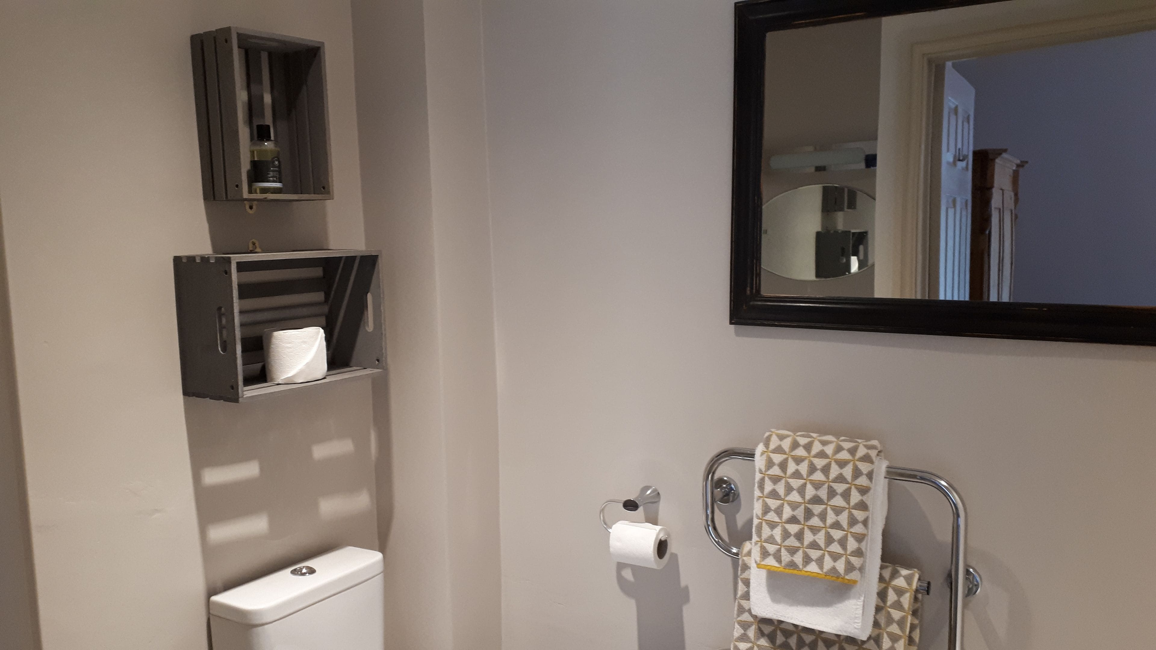 A bathroom with a toilet, a towel rack with a patterned towel, a mirror, and a shelf with a bottle and a tissue box.