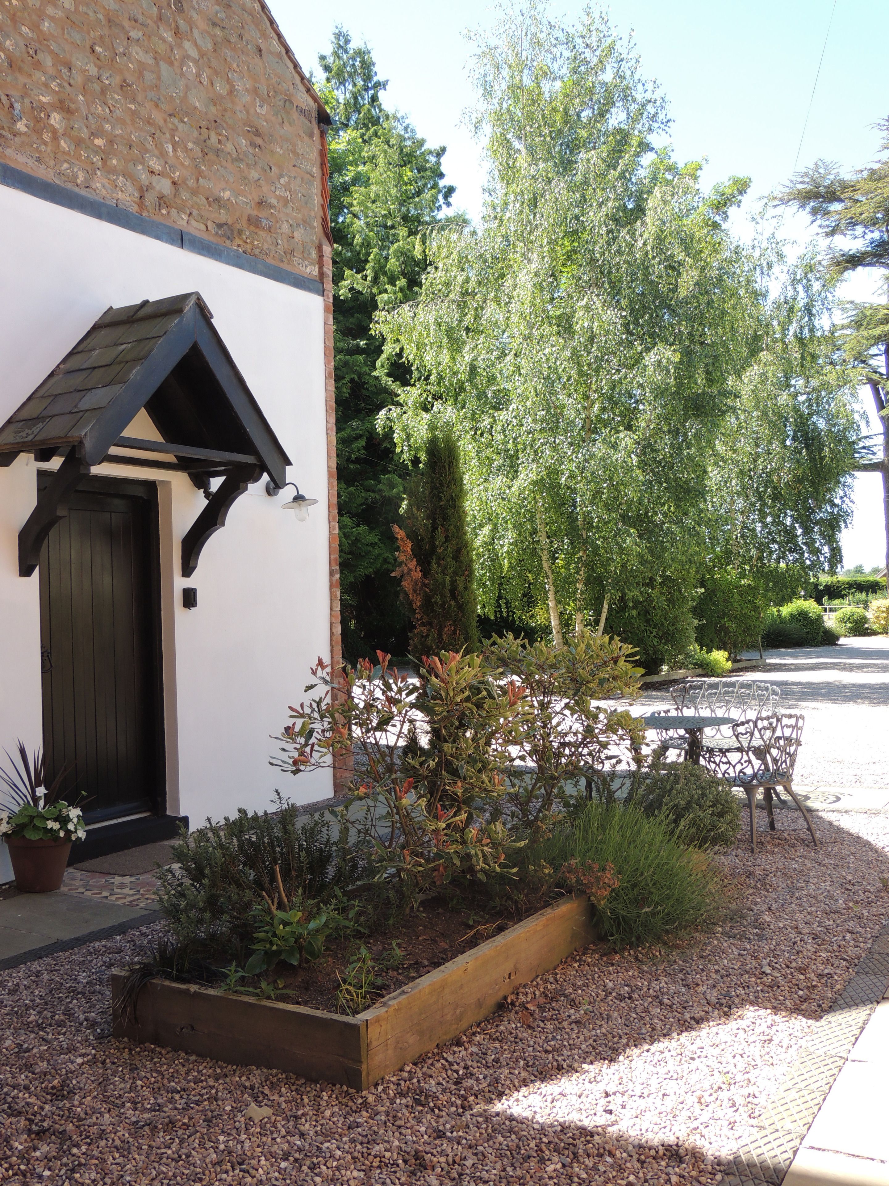 A building with a sloped roof, a wooden door, and a small porch. There is a garden with gravel, plants, and a metal table and chairs.