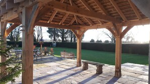 Una pérgola de madera con suelo de piedra y mobiliario exterior, incluyendo un banco y sillas, con vista a un césped bien cuidado.