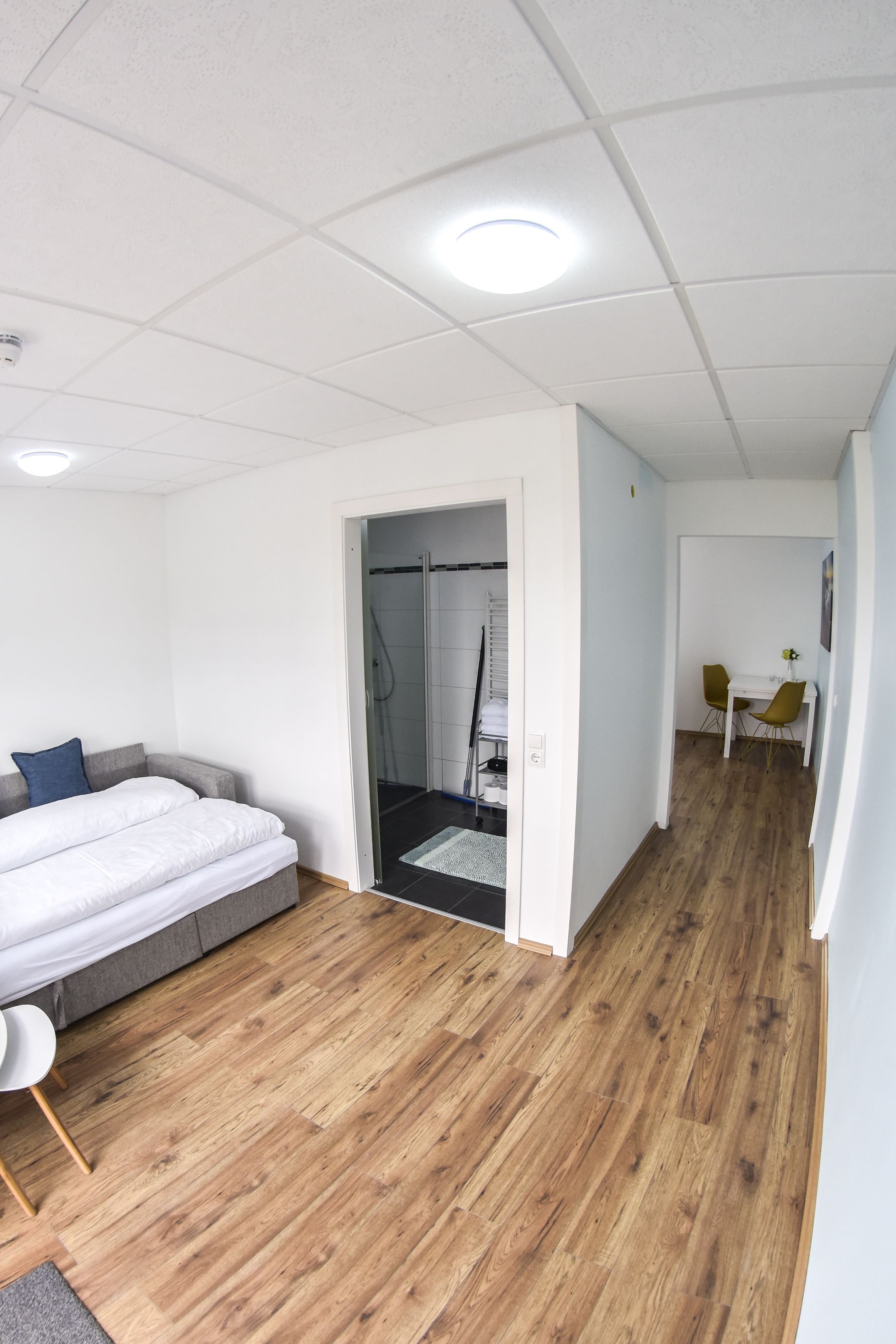 Quarto de hotel moderno com piso de madeira, uma cama com cabeceira cinza, uma pequena mesa branca e uma cadeira amarela.