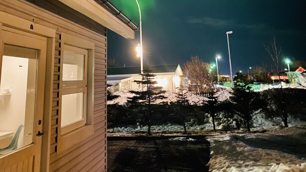 Un edificio con entrada y ventanas visibles, suelo cubierto de nieve y vista al cielo nocturno con auroras verdes.