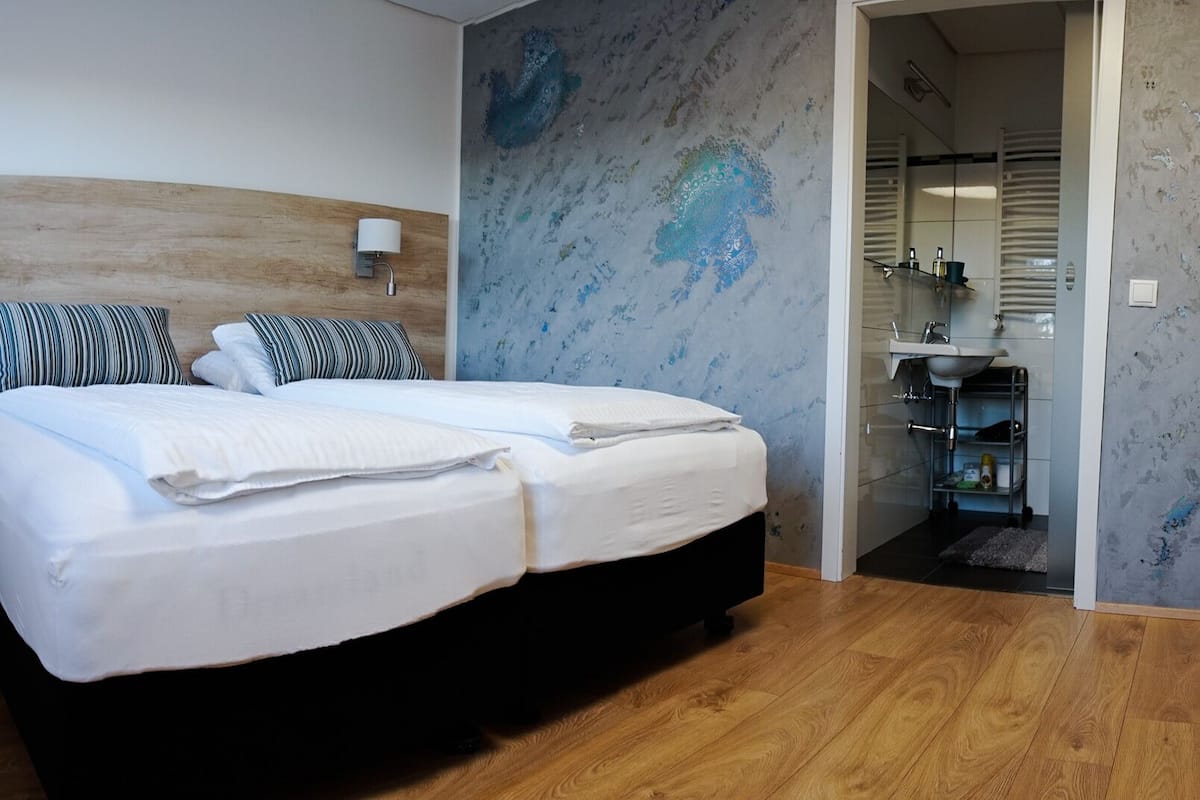 Un dormitorio con cama, mesita de noche, una silla y un baño visible a través de una puerta abierta.