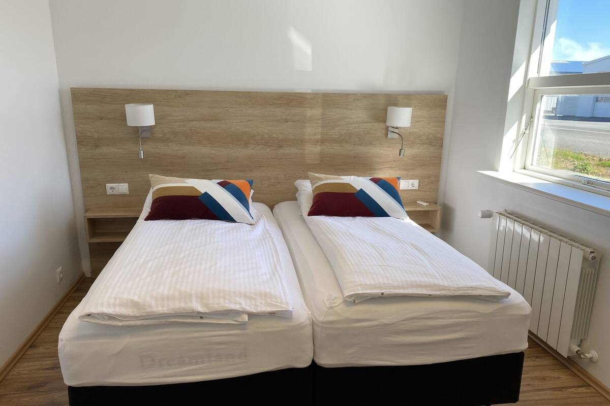 Habitación doble con cama y cabecera de madera, lámparas de noche y ventana con vistas.