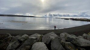 Una playa con rocas grandes al frente y una persona de pie en el agua.