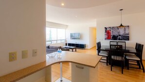 A modern living room with a curved kitchen counter, a dining area with chairs, a sofa, and a TV mounted on the wall. - Capitalia | Renovated 2BR in Santa Fe’s Heart (Ciudad de México)