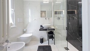 Un baño moderno con inodoro blanco, un bidé, un espejo grande, un lavabo de mármol negro y una zona de ducha con paredes de vidrio.