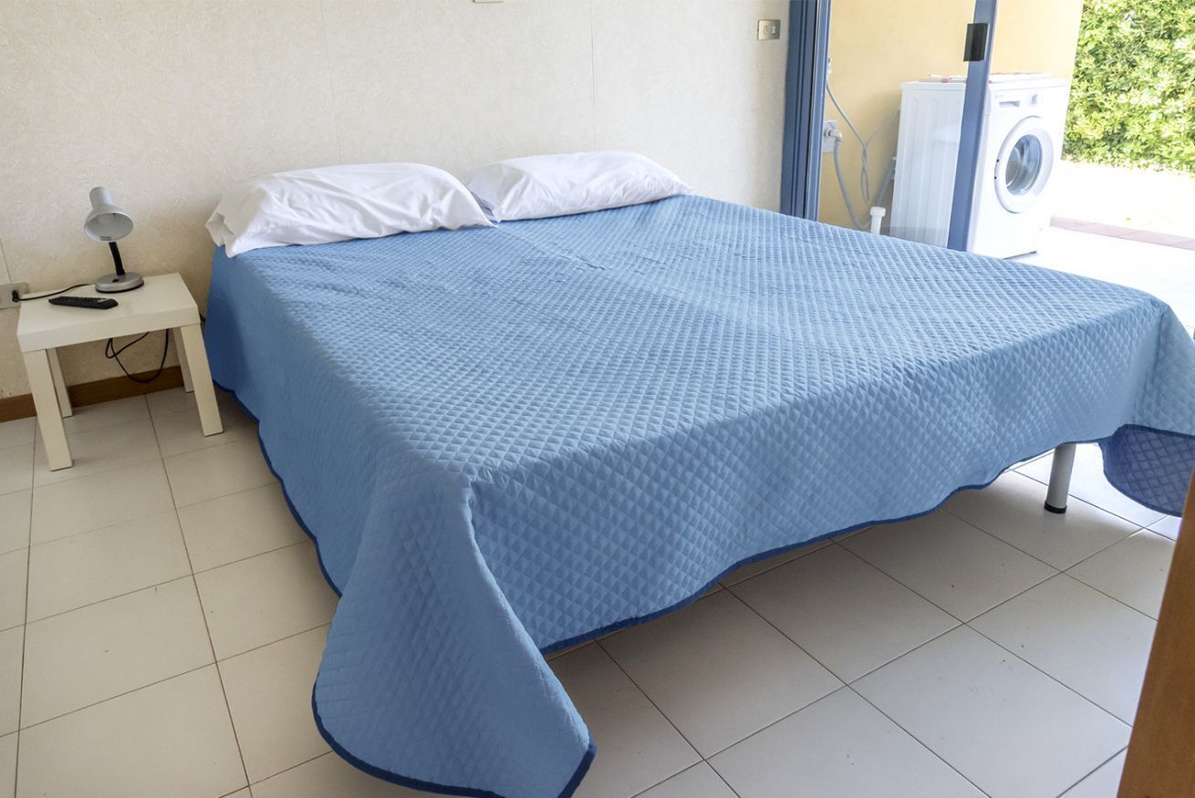 A bed with a blue quilt, a white pillow, a nightstand with a lamp, and a washing machine.