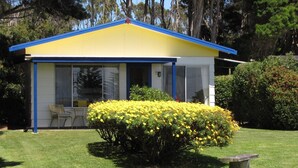 Un edificio amarillo de una sola planta con techo azul, un porche cubierto con mesa y sillas, y un césped cuidado con arbustos florecientes amarillos.
